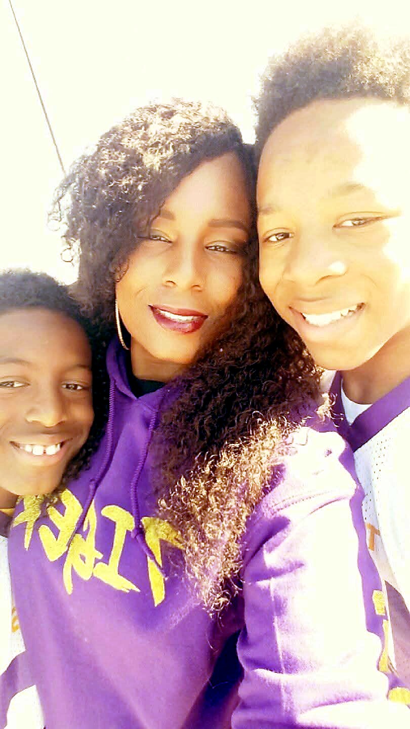 Three smiling people posing for a sunny selfie outdoors: a woman in the middle with two children on either side, all wearing purple clothing.