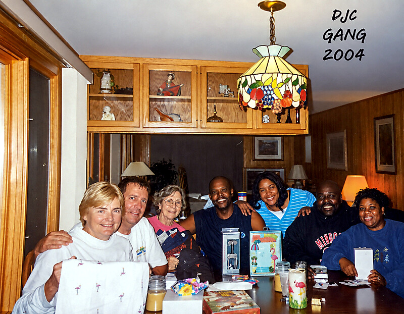 Group of eight adults posing for a casual photo around a dining table with gifts, in a wood-paneled living room; warm, friendly gathering. Text 'DJC GANG 2004' appears in the upper right.