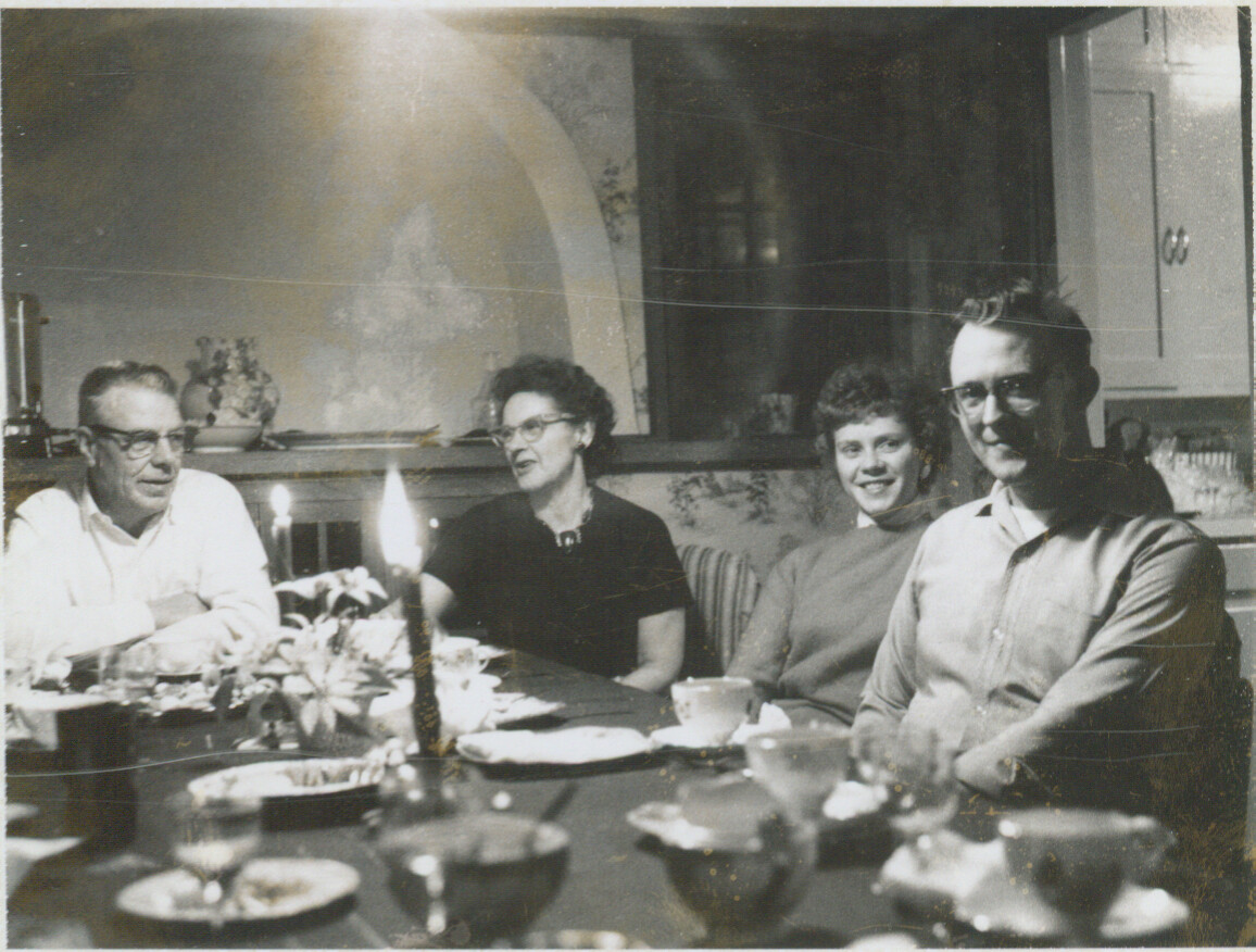 Black-and-white photo of four adults seated around a dining table with candles and dishes, smiling in a home kitchen/dining room.