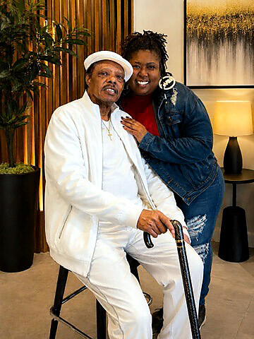 Older man in a white suit and hat sits on a stool with a cane, smiling, as a woman in a denim jacket stands beside him indoors with plants in the background.