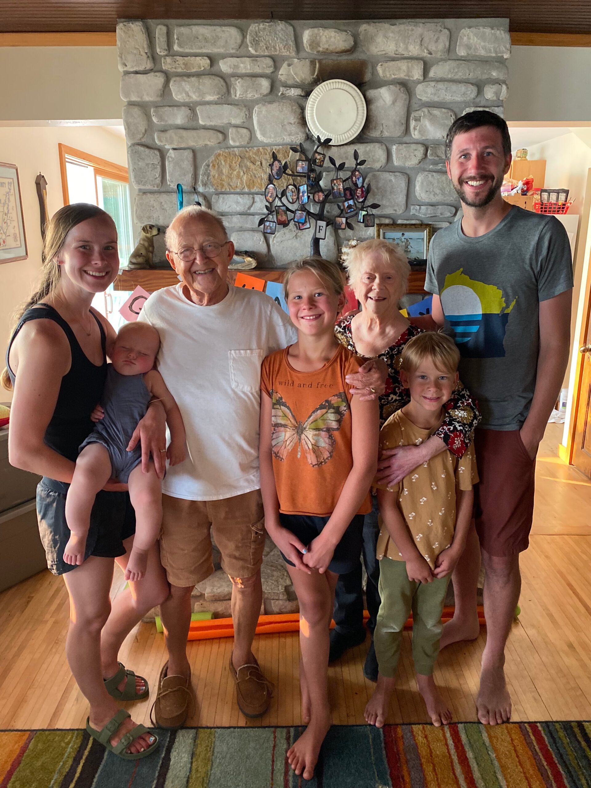 Multi-generational family posing indoors in front of a stone fireplace, smiling at the camera.