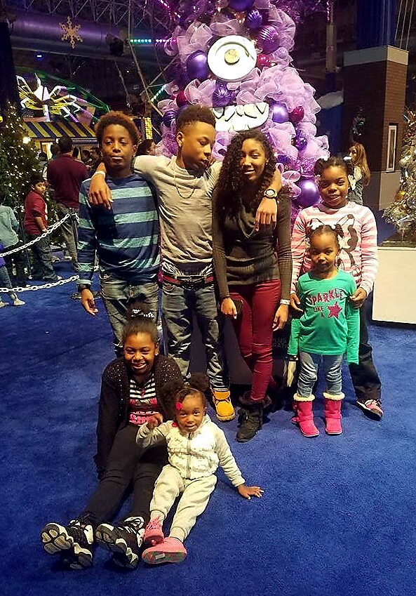 A group of eight children posing together inside a festive venue with a purple decorative tree in the background, smiling and posing for the camera.