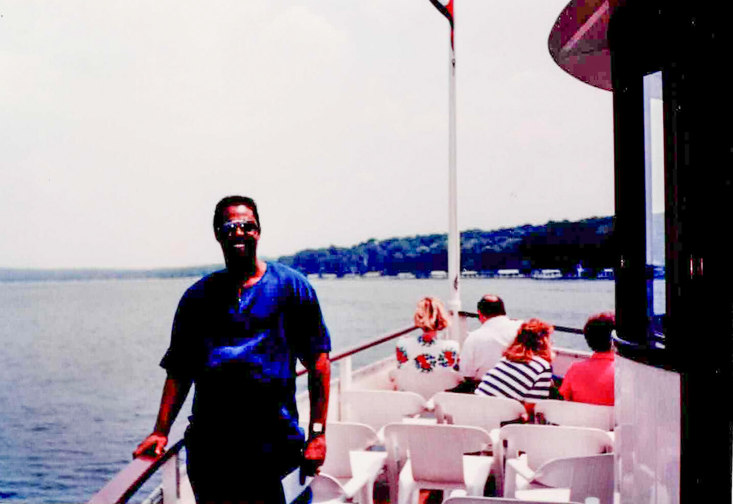 Man standing at the railing of a boat deck, smiling, with passengers seated behind him on white chairs by the water.