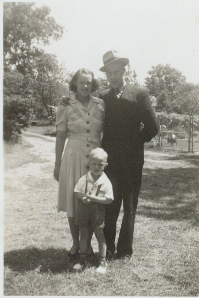 A smiling woman in a light dress stands with a man in a dark suit and hat, joined by a small child, outdoors in a sunny park setting.