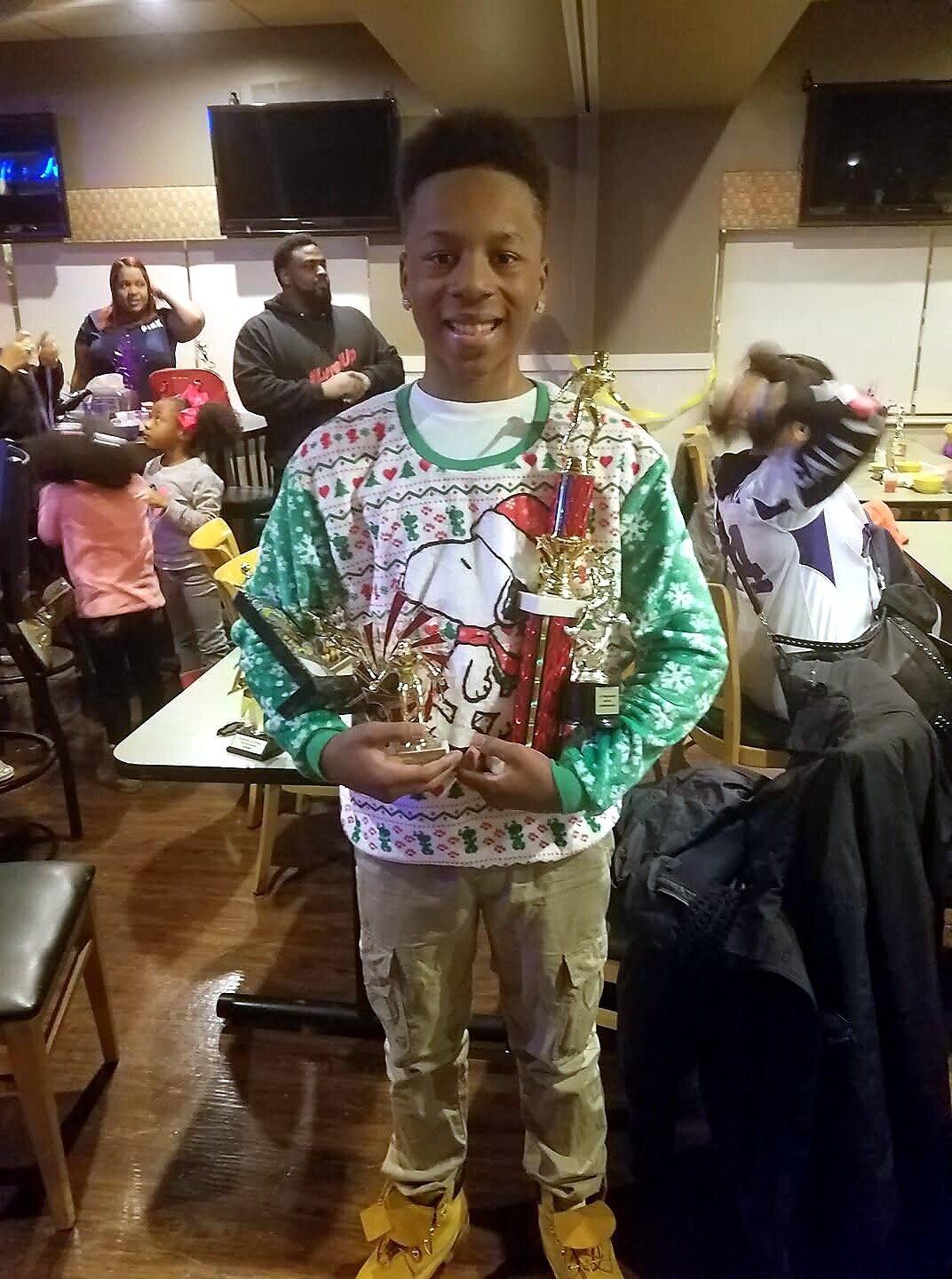 Young man in a festive Christmas sweater holding several trophies at a party, smiling at the camera in a diner or lounge setting with kids in the background.