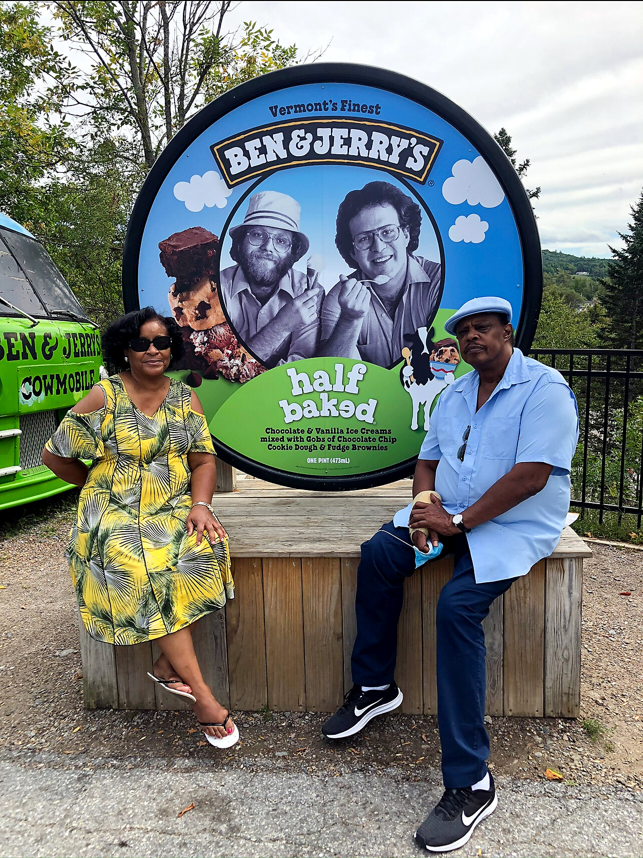 Two people sit on a wooden platform in front of a Ben & Jerry's sign featuring half baked flavor artwork outdoors on a cloudy day.