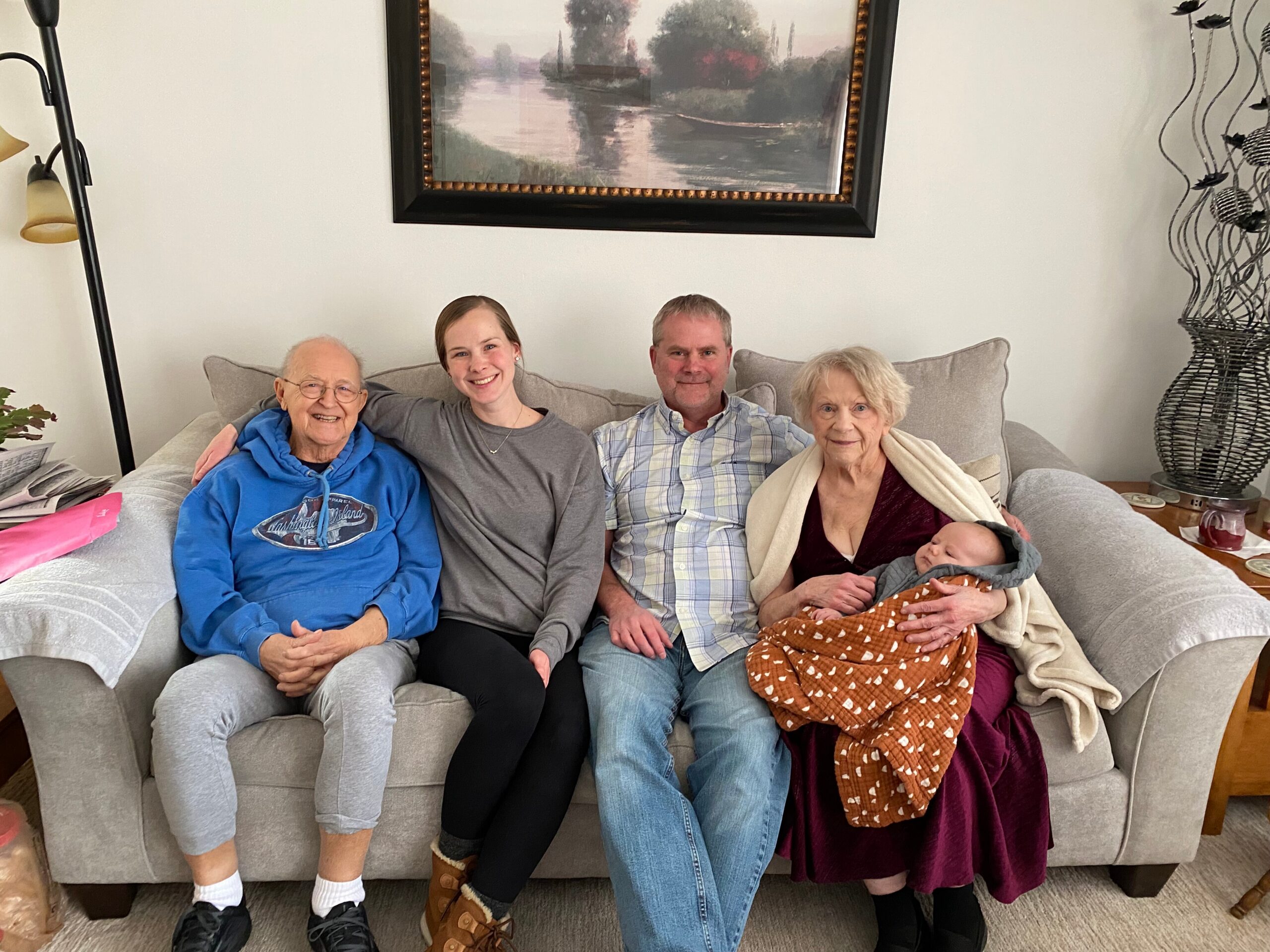 A multigenerational family sits together on a gray sofa in a living room, from left: an elderly man in a blue hoodie, a younger woman, a man in a plaid shirt, an elderly woman with a baby tucked in a brown blanket on her lap.