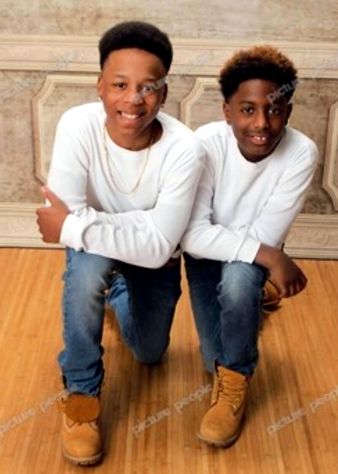 Two men kneeling on a wood floor, wearing white shirts, blue jeans, and tan boots, smiling at the camera.