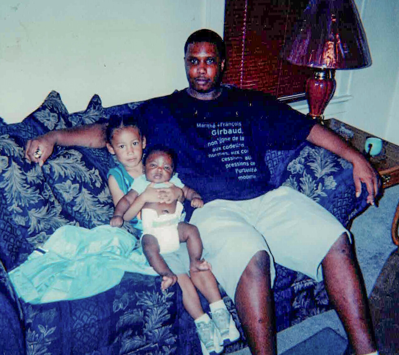 A man sits on a patterned sofa with two young children, one held on his lap and another beside him, in a living room setting.