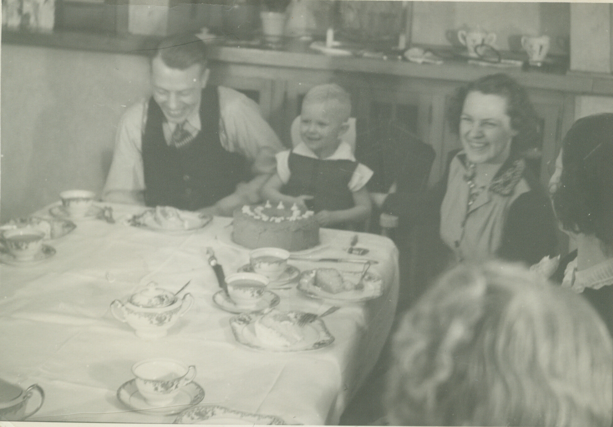 Three people smile at a birthday table with a frosted cake and tea cups in front of them.