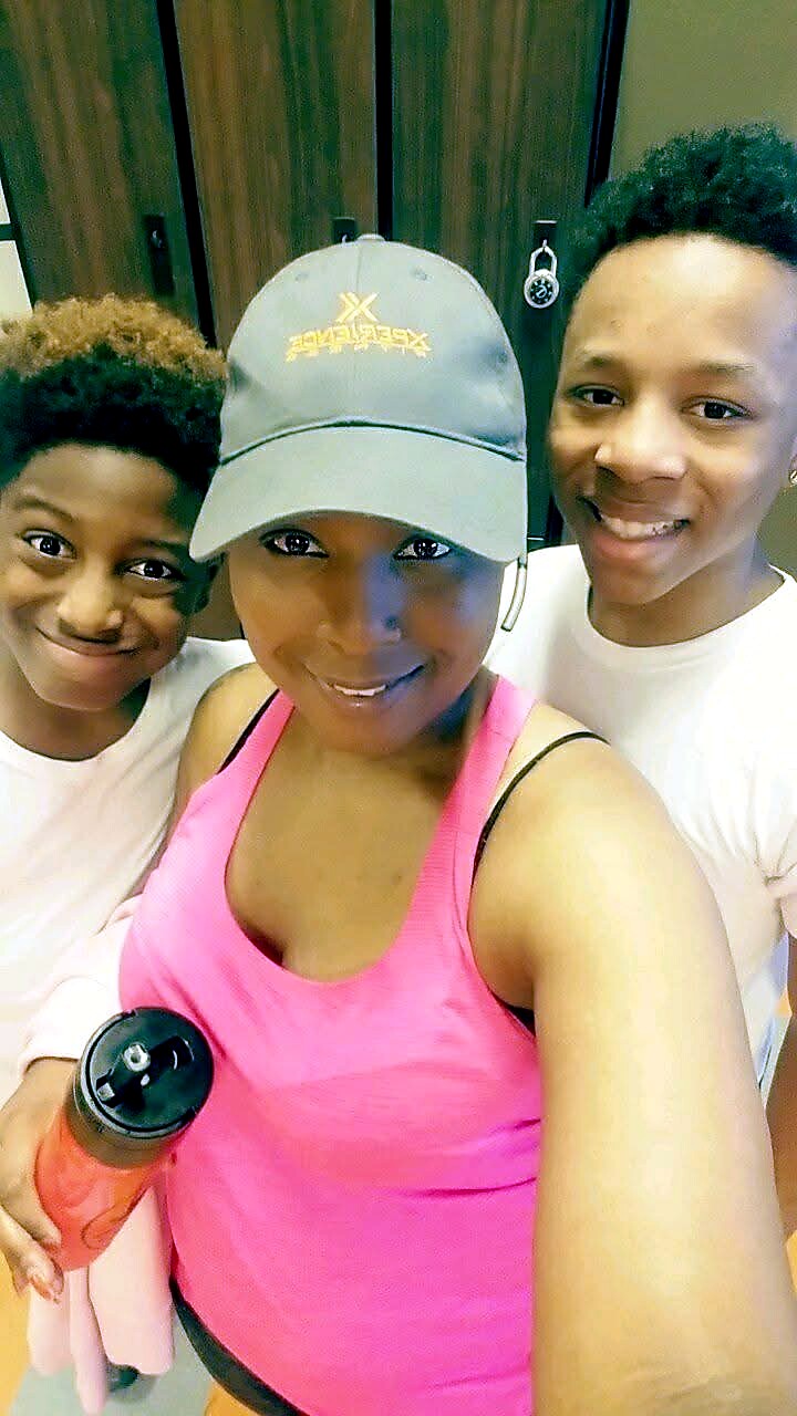 Three people posing for a selfie in a locker room; woman in a pink gym top and gray cap in the center, flanked by two boys in white shirts.