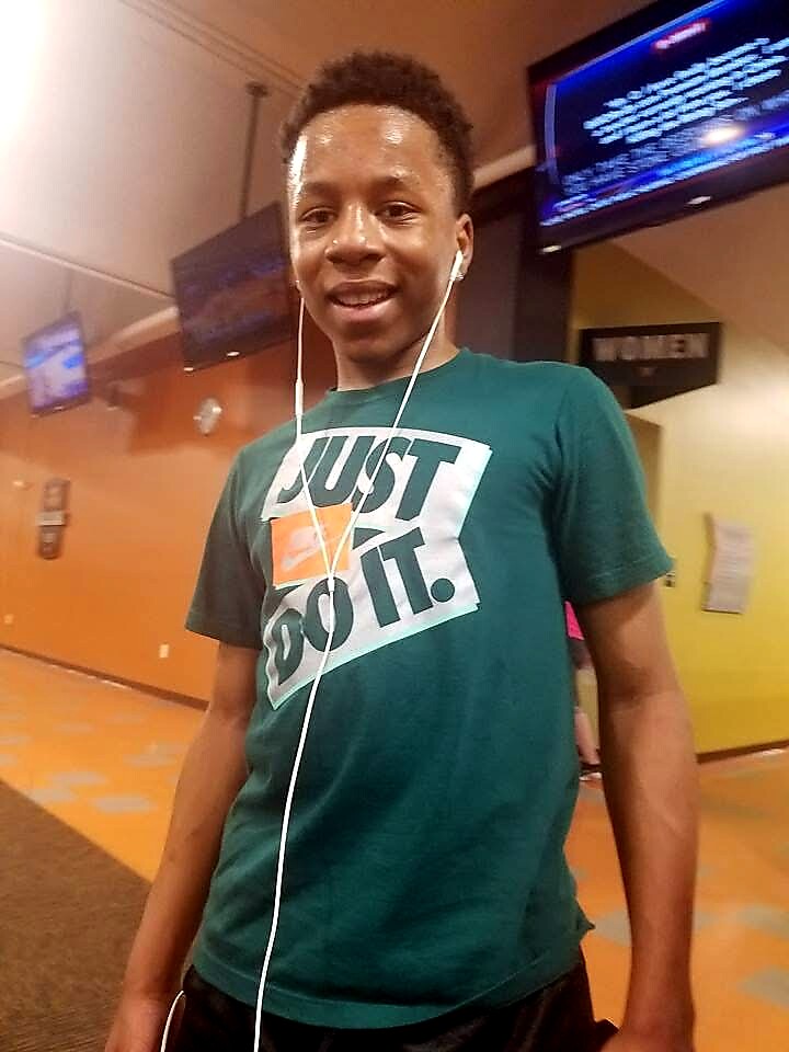Young man in a teal Nike-style shirt with the slogan 'Just Do It.' wearing white earphones, standing in a gym or hallway and smiling at the camera, with screens visible in the background.