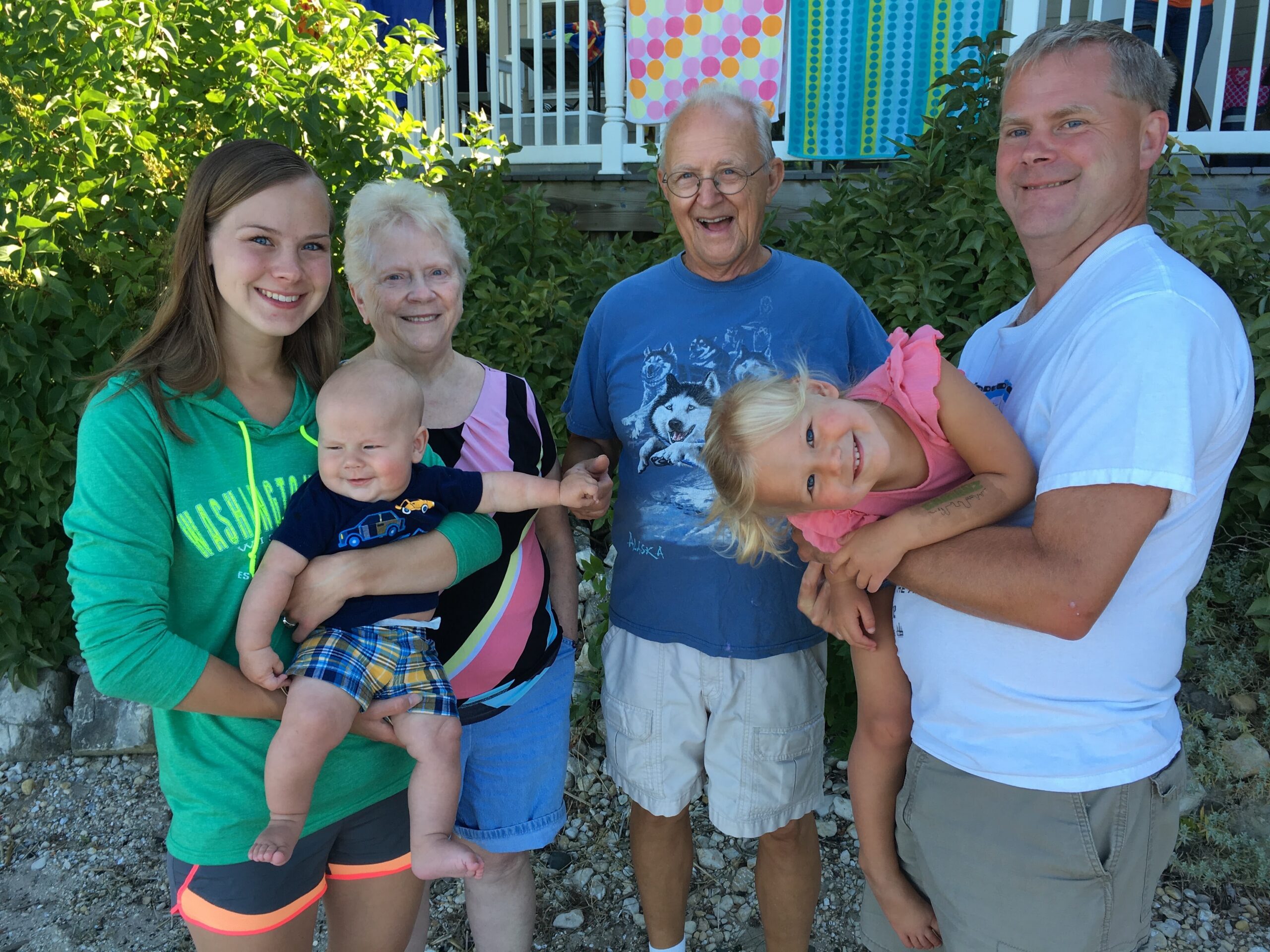 Smiling group outdoors: young woman in green hoodie holding a baby, an older woman beside her, a man in a blue shirt, and a man in a white shirt holding a young girl leaning to the side.
