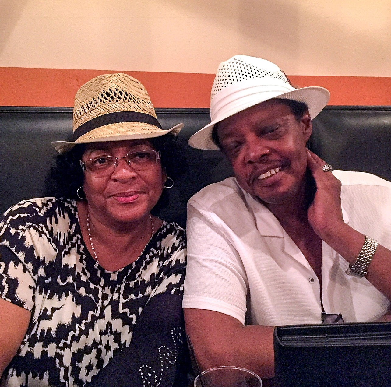 Two smiling older adults sit together in a restaurant booth; woman wears a straw hat and patterned top, man wears a white hat and light shirt with jewelry nearby.