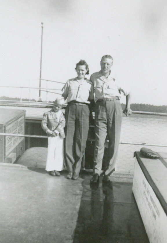 A couple in uniformed shirts and high-waisted trousers stand arm in arm on a rooftop deck with a young girl in a dress beside them; a railing and water are in the background.