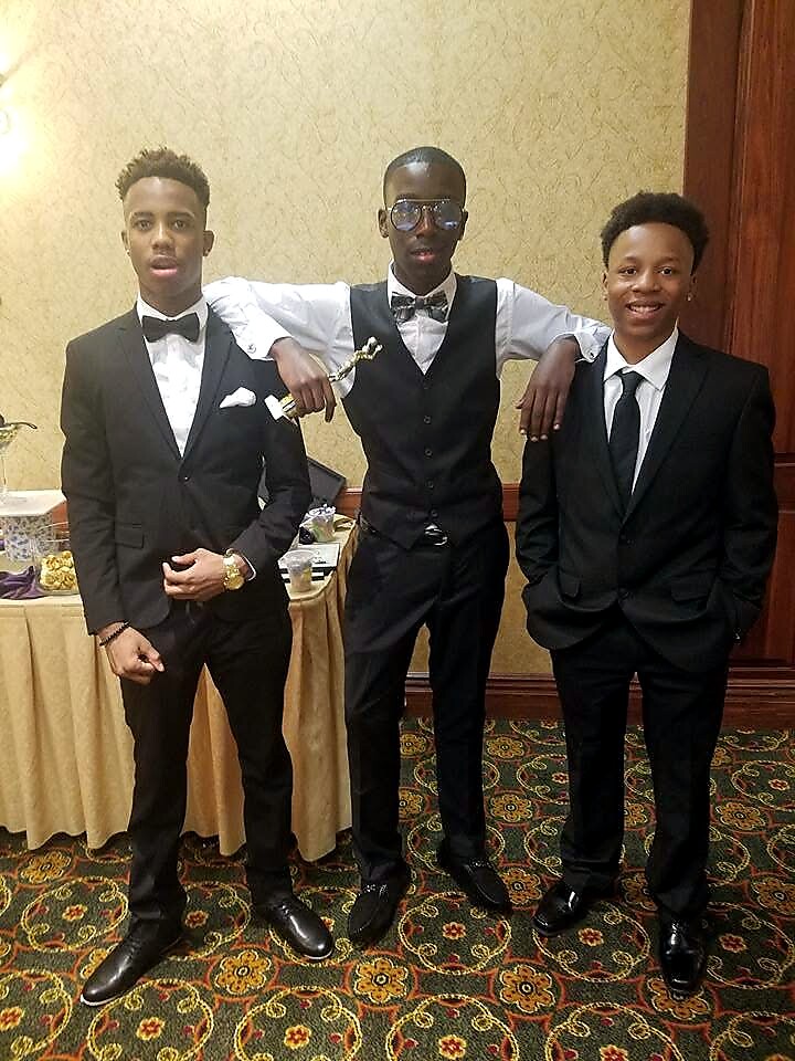 Three young men in formal black suits posing at a banquet hall; the middle man wears sunglasses and holds a small trophy or prop while flanked by two friends.