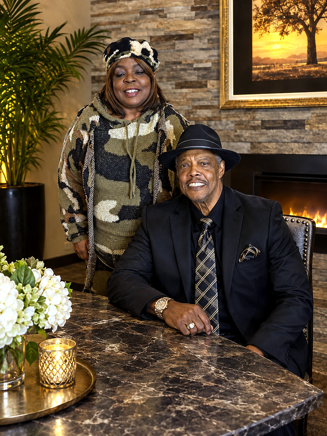 A smiling man in a black suit and hat sits at a marble table, while a woman in camo-patterned clothing stands behind him in a cozy, candlelit room.