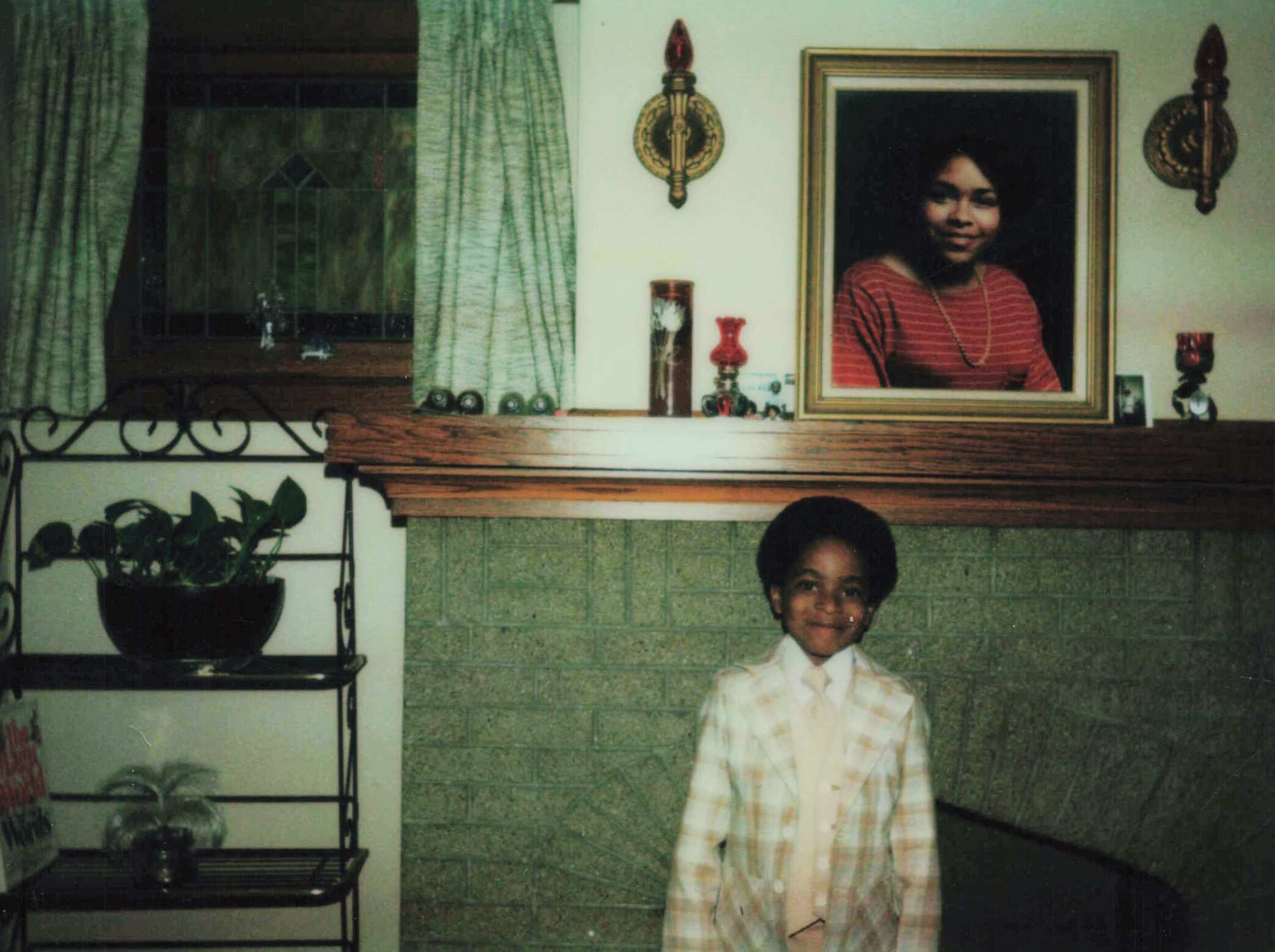 Child standing indoors in front of a fireplace mantel, wearing a light plaid dress, with a framed portrait of a girl on the mantel and decorative items nearby.