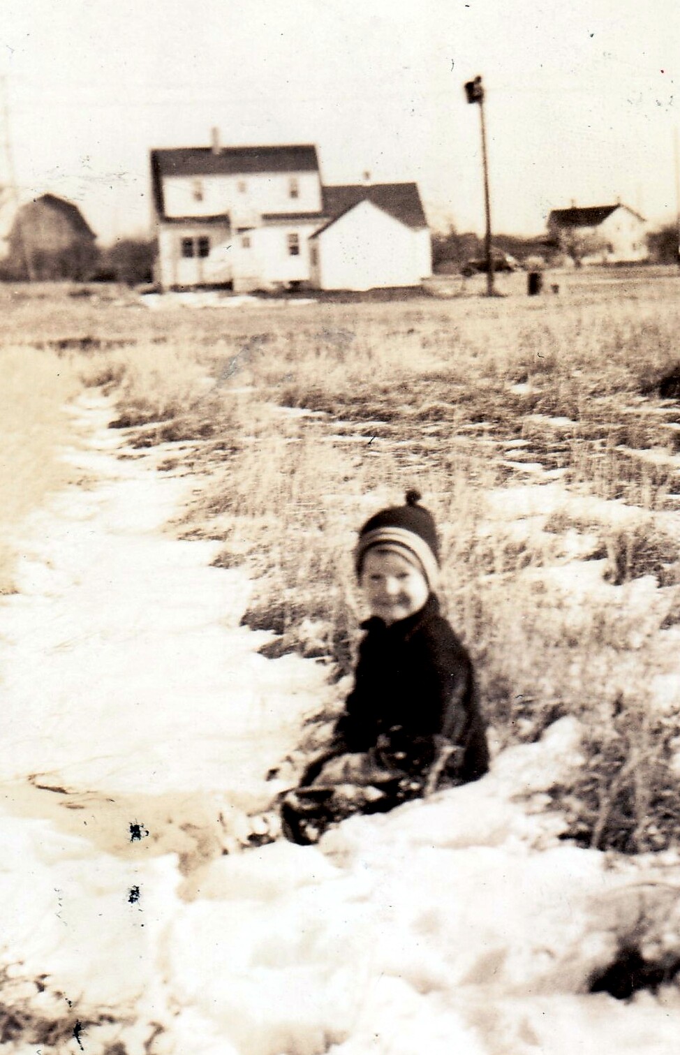 Child in a dark coat and striped hat kneels in a snowy field, facing the camera, with houses and a streetlight in the distance.