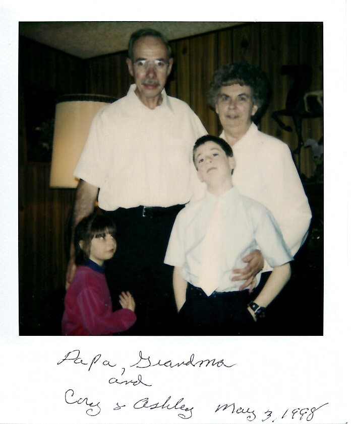 Portrait of a grandfather, grandmother, and two children posing in a living room; handwritten caption below reads 'Papa, Grandma and Cy & Ashley May 3, 1988'.