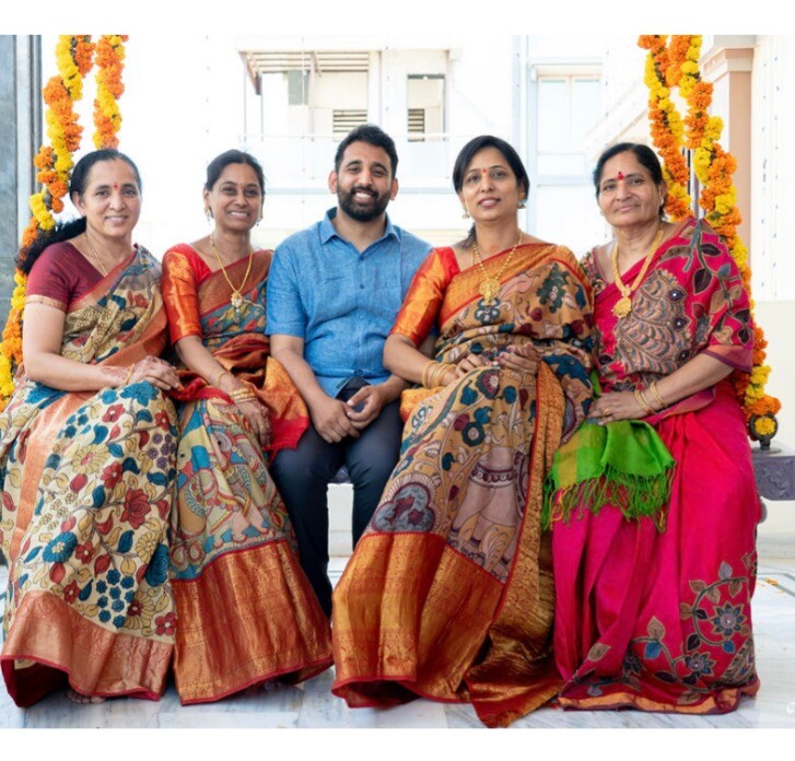 Man in a blue shirt sits between four women in colorful sarees, posing for a family photo at a festival with marigold garlands.