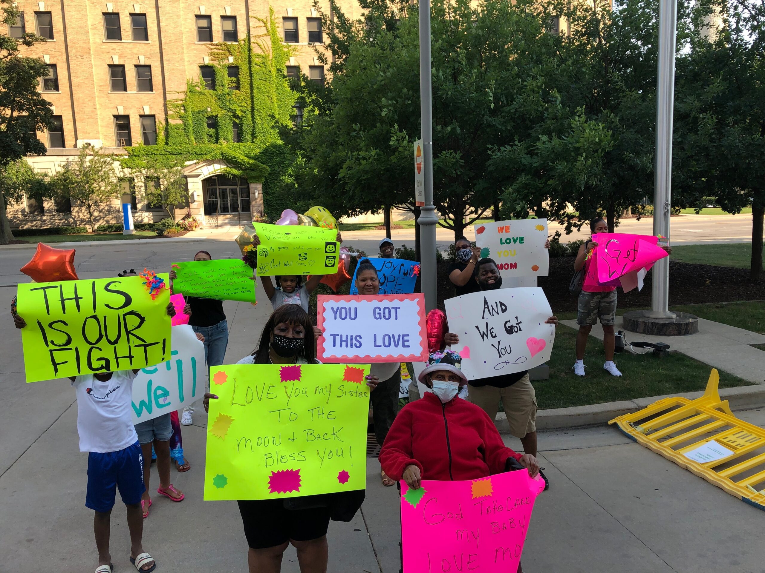 Group of protesters, including children and adults, holding neon signs with messages of love and support on a sunny campus sidewalk near a brick building with ivy.