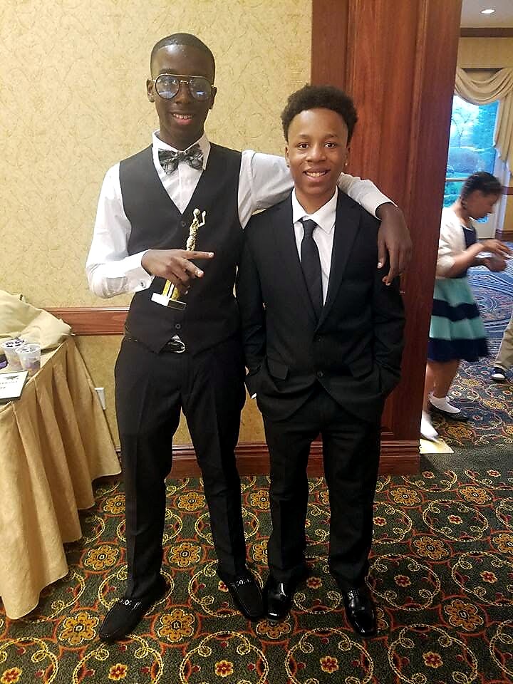 Two young men in formal attire pose together indoors; one holds a trophy and wears glasses and a bow tie while the other wears a black suit and tie.
