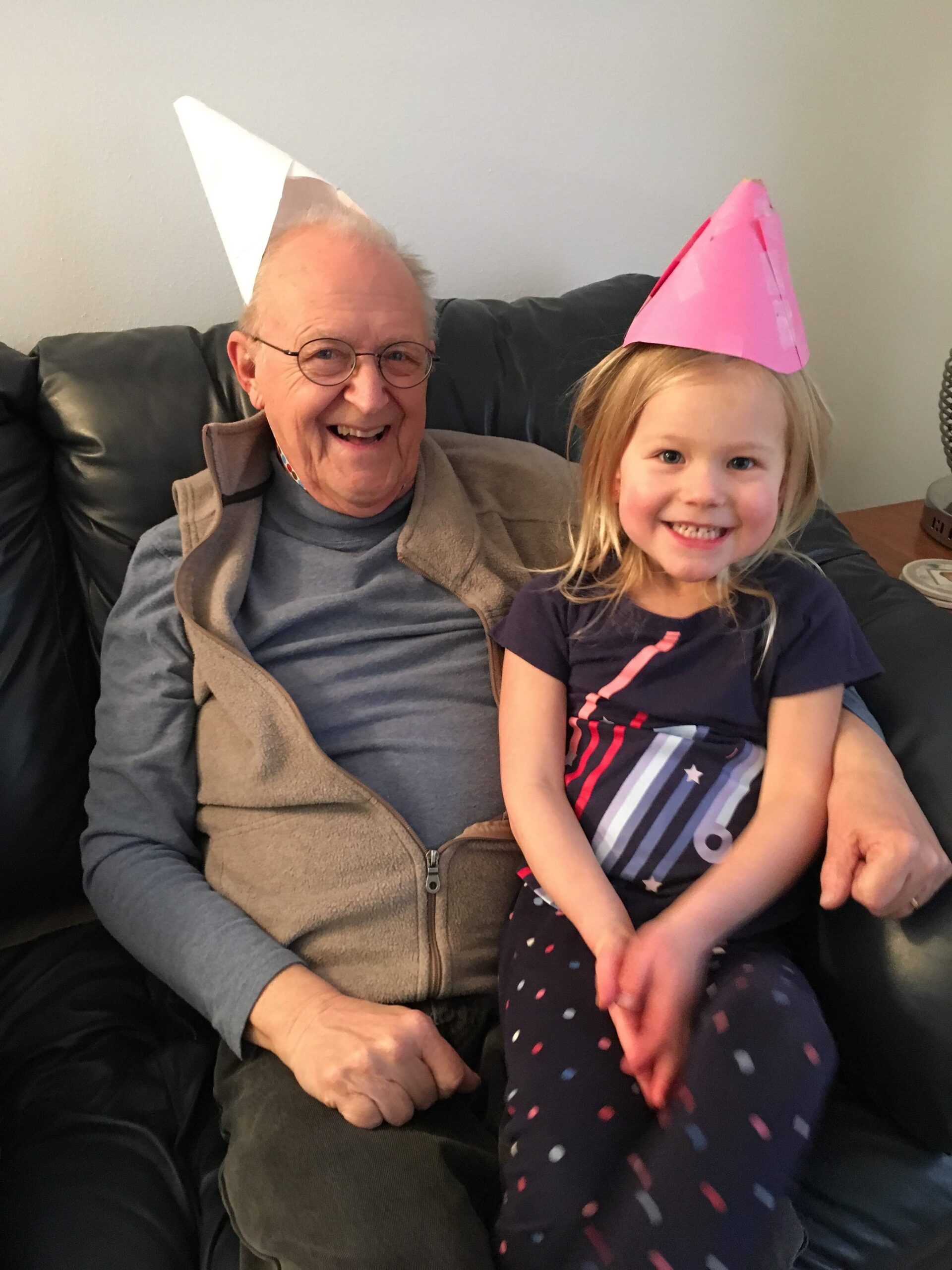 Smiling older man and young girl wearing party hats on a couch in a living room