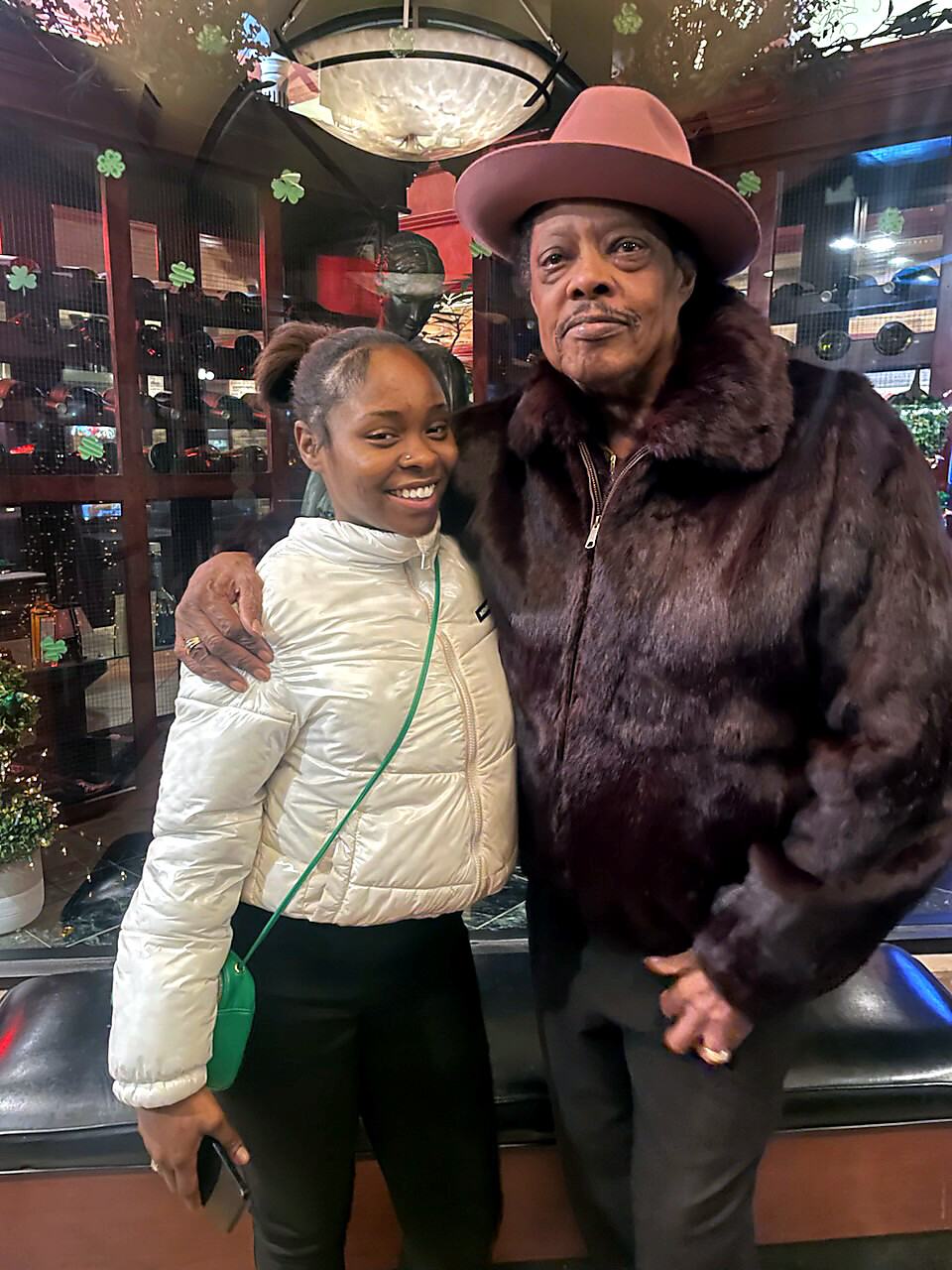 Two women pose together smiling; one in a white puffer jacket with a green crossbody bag, the other in a dark fur coat and pink hat in a storefront window display.