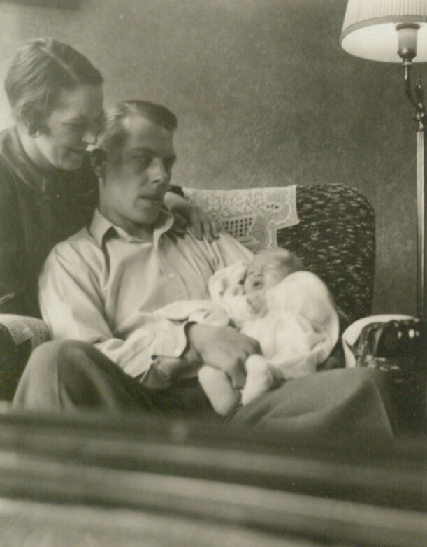 Father seated with a baby on his lap as a woman leans in from the left, smiling in a cozy living room.