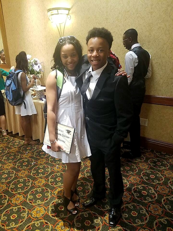 Two teens in formal attire pose together at a banquet, the girl in a white dress holding a program and the boy in a black suit smiling.