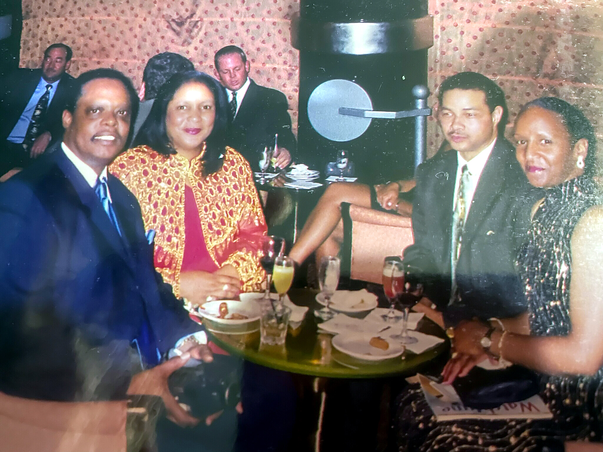 Group of adults in formal attire at a celebration around a table with drinks and cake.