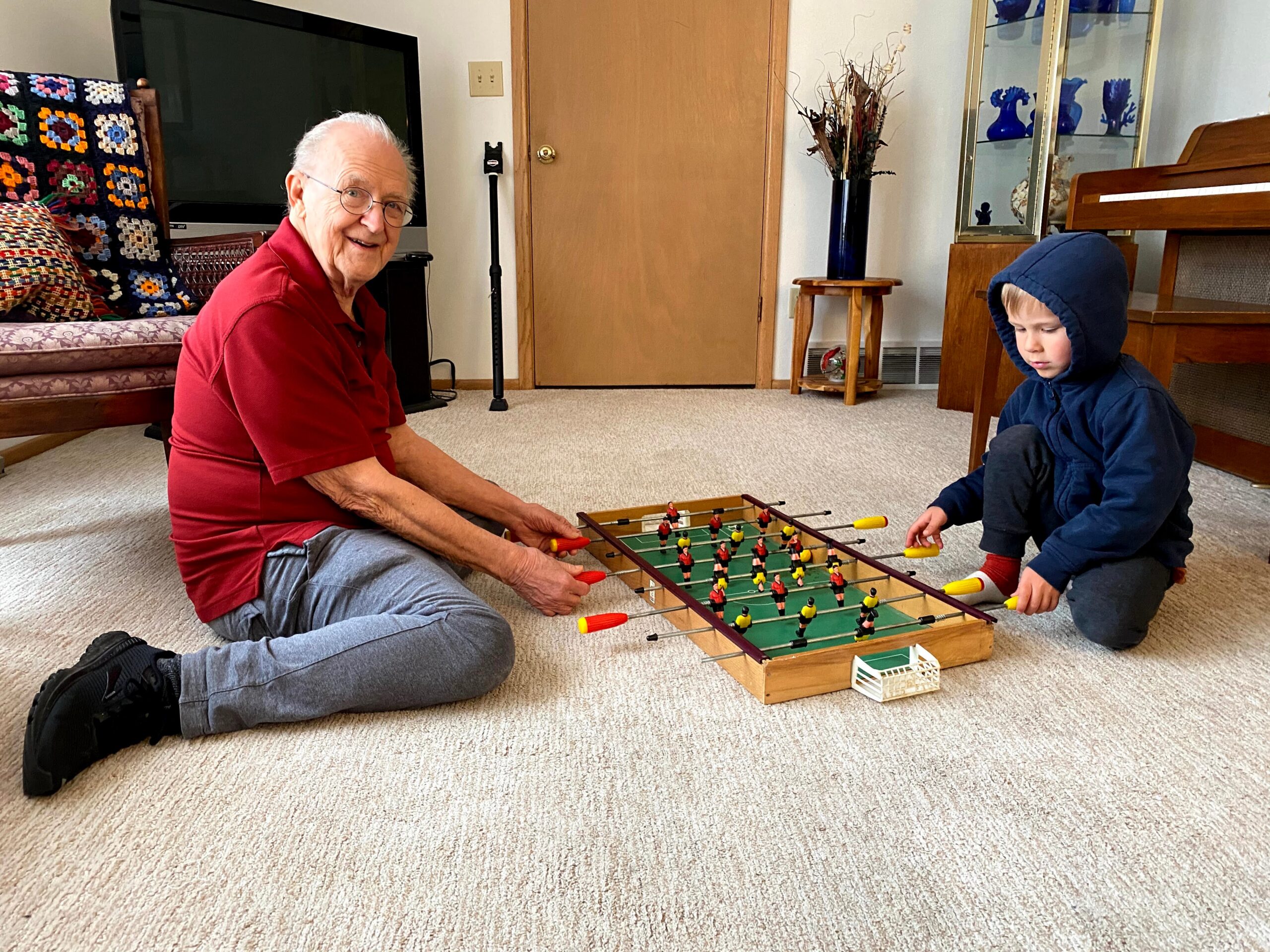 Older man in a red shirt and a child in a blue hooded jacket playing foosball on a wooden table in a living room.