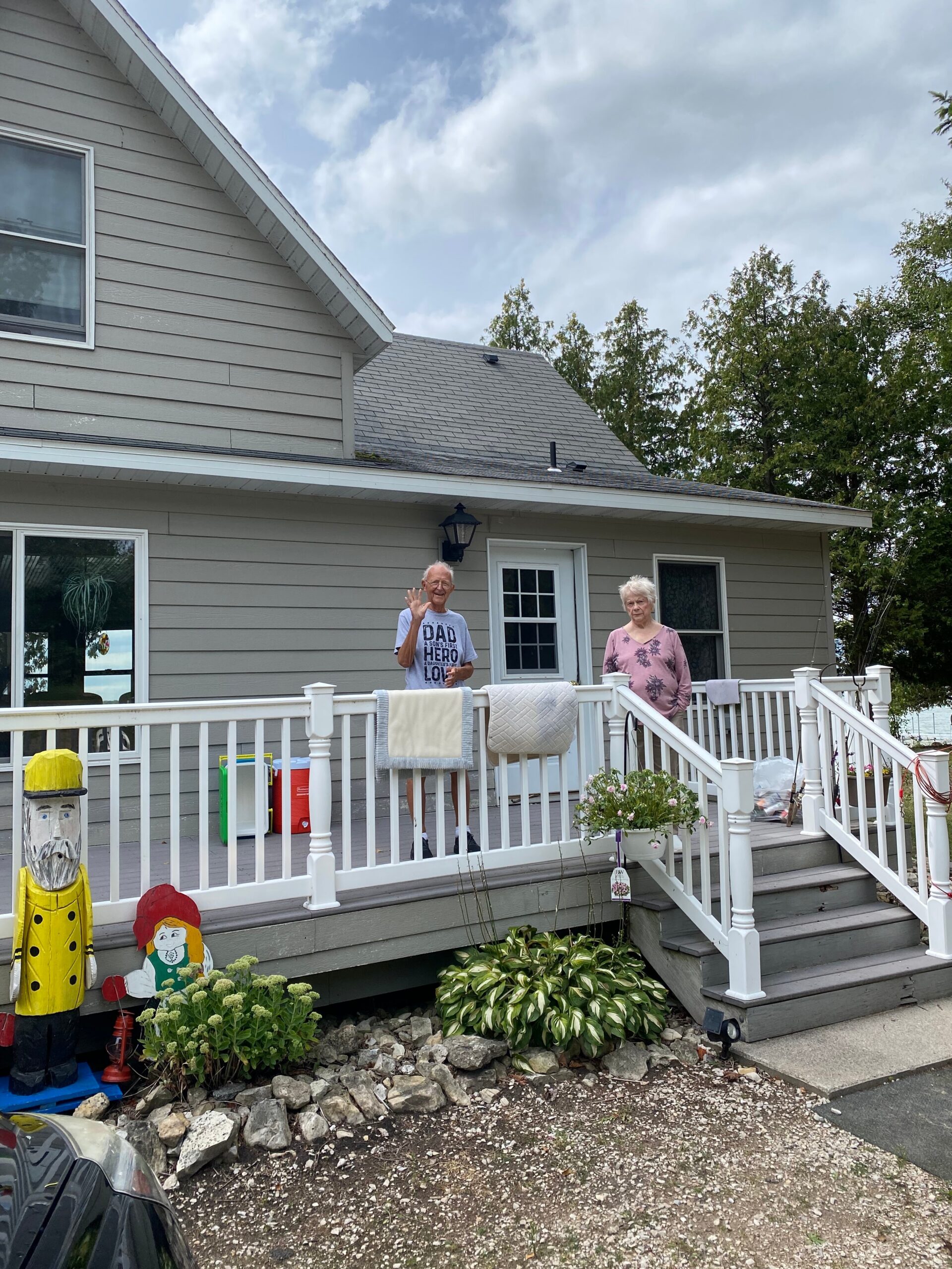 Two elderly people stand on a white-railed front porch of a gray house, smiling and waving to the camera.
