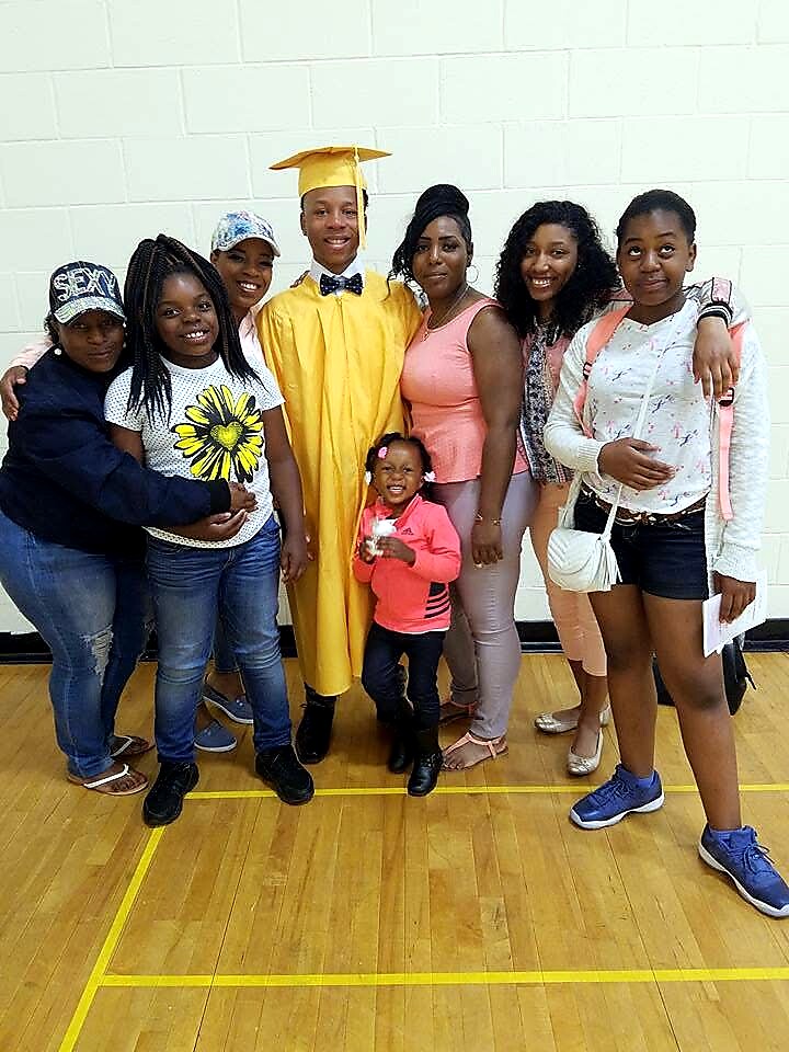 A graduate in a yellow cap and gown poses with seven family and friends in a gym, all smiling for the photo.