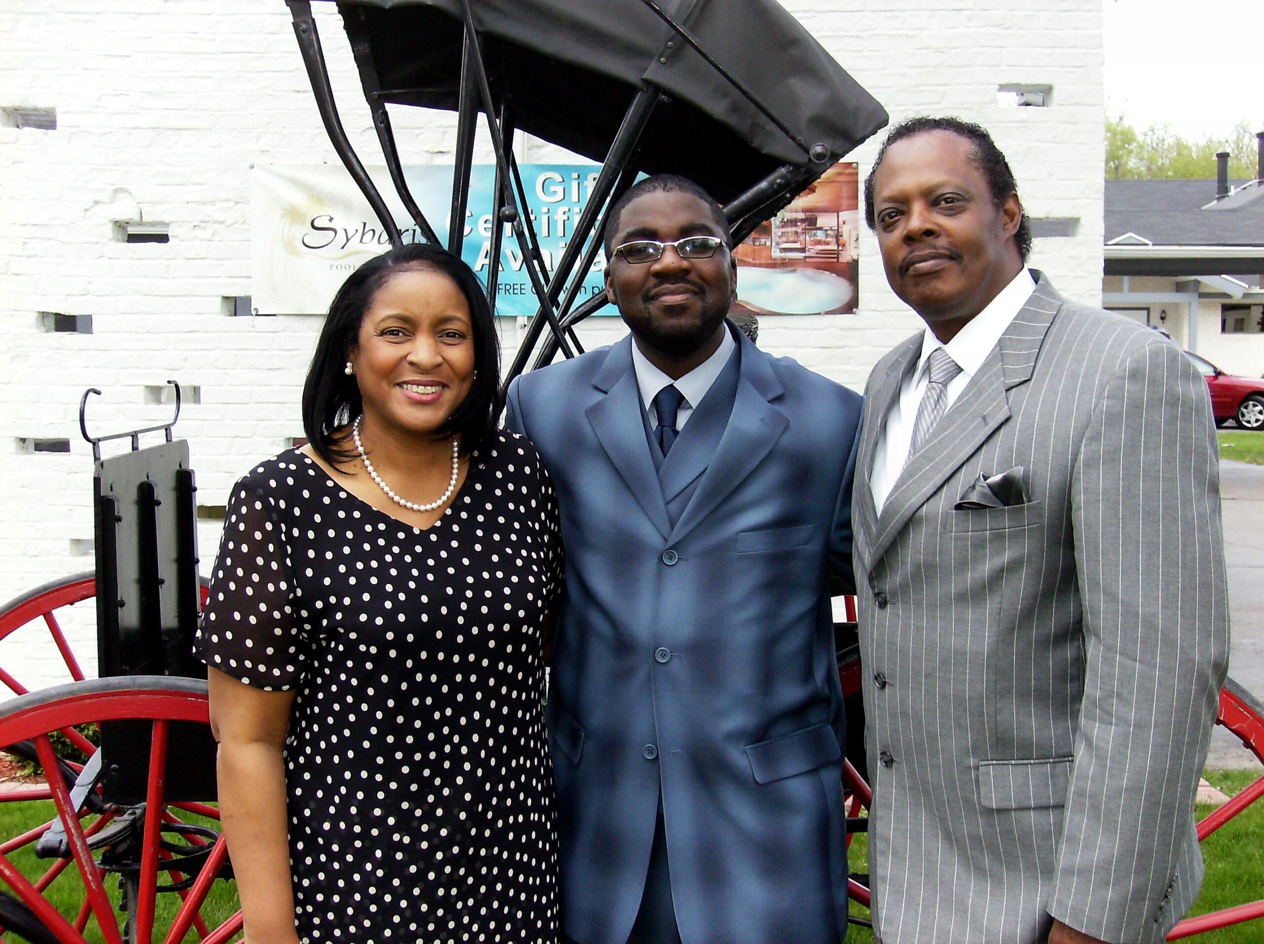 Three adults in business attire posing for a photo outdoors in front of a white brick wall with a red wagon wheel visible to the left. From left to right: a woman in a black polka-dot dress with a pearl necklace, a man in a blue suit and glasses, and a man in a gray pinstripe suit.