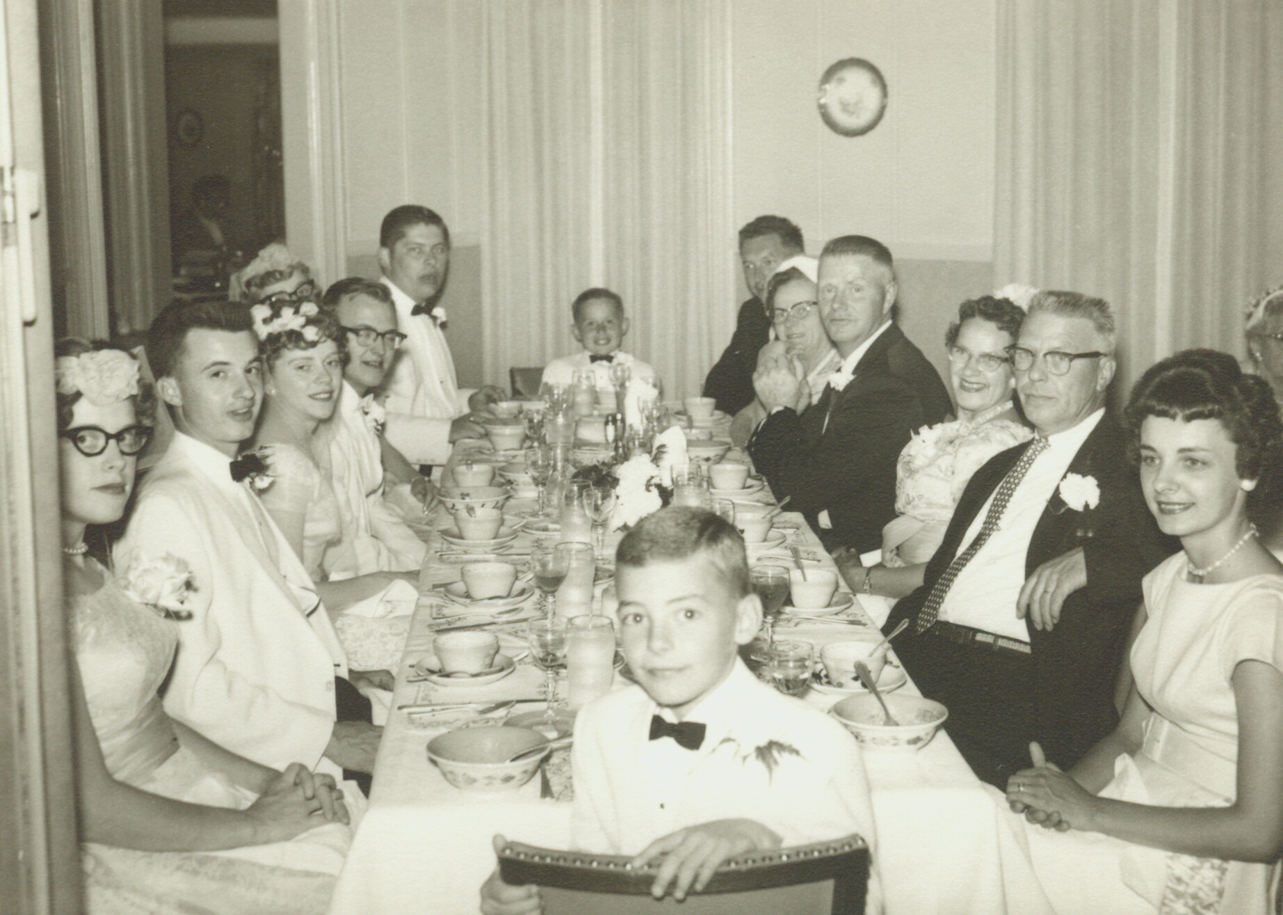 Wedding reception with guests in formal attire seated along a long dinner table, showing bowls, cups, and a floral centerpiece.
