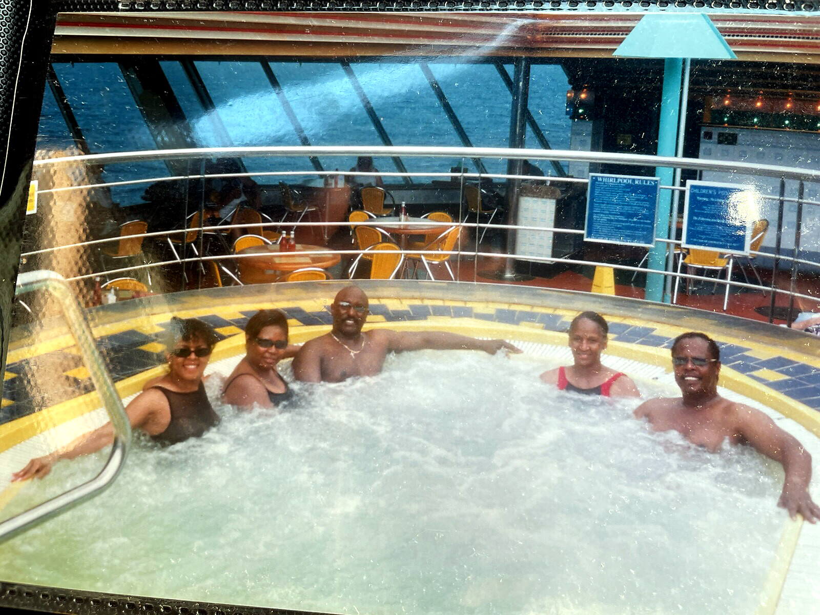 Six adults sit in a circular indoor jacuzzi on a cruise ship, smiling at the camera.