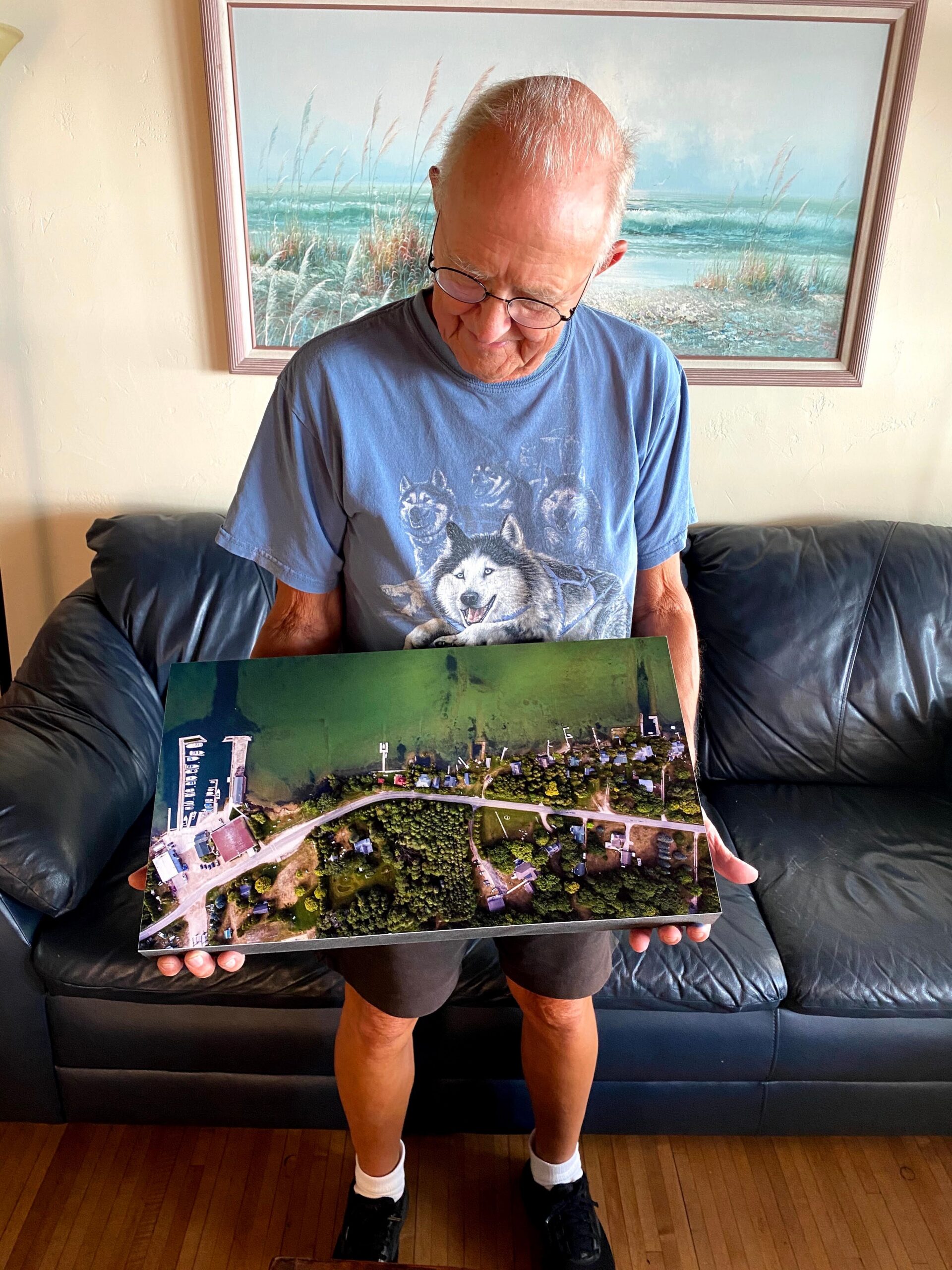Older man with glasses in a blue wolf shirt looks down at a large aerial photograph on his hands.