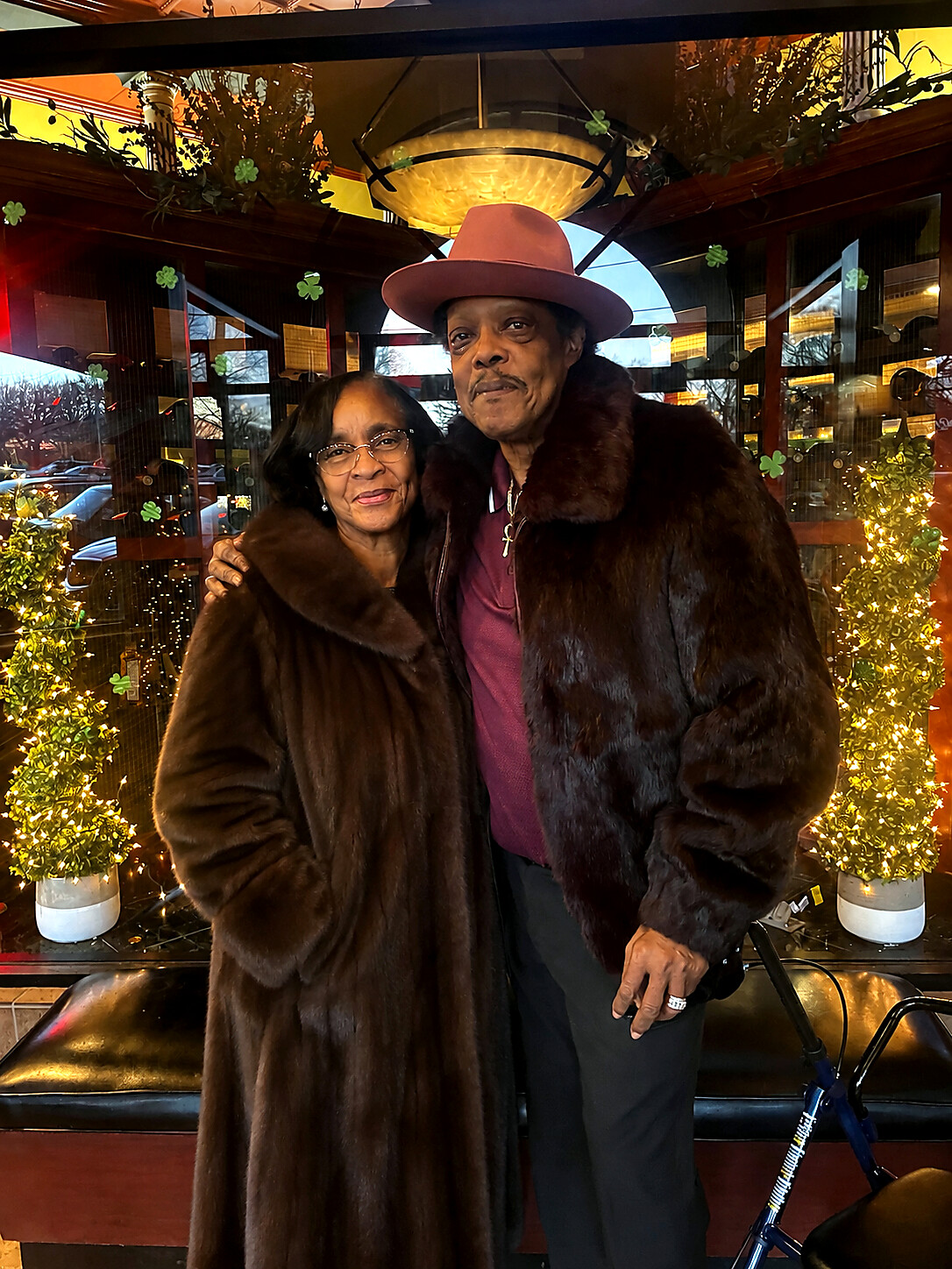 An elderly couple poses for a photo in a warmly lit, festive setting, both wearing dark fur coats; the man wears a pink hat and a cross necklace.