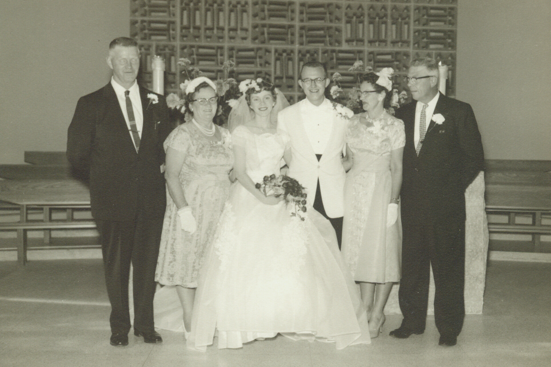 Bride and groom stand with their parents in a church wedding photo, all smiling in formal attire.