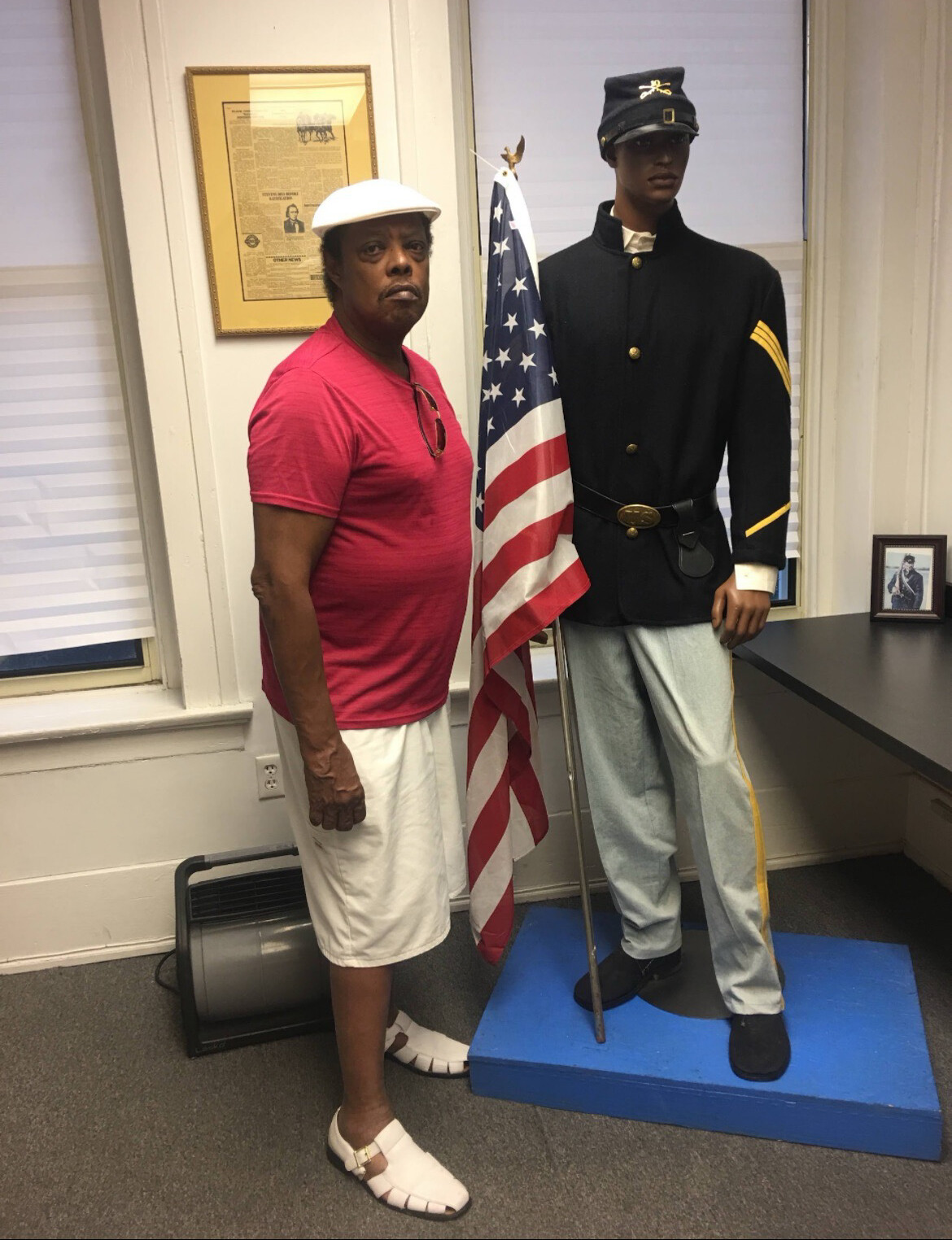 Man in a red shirt and white cap posing beside a military mannequin holding the American flag in a display room.
