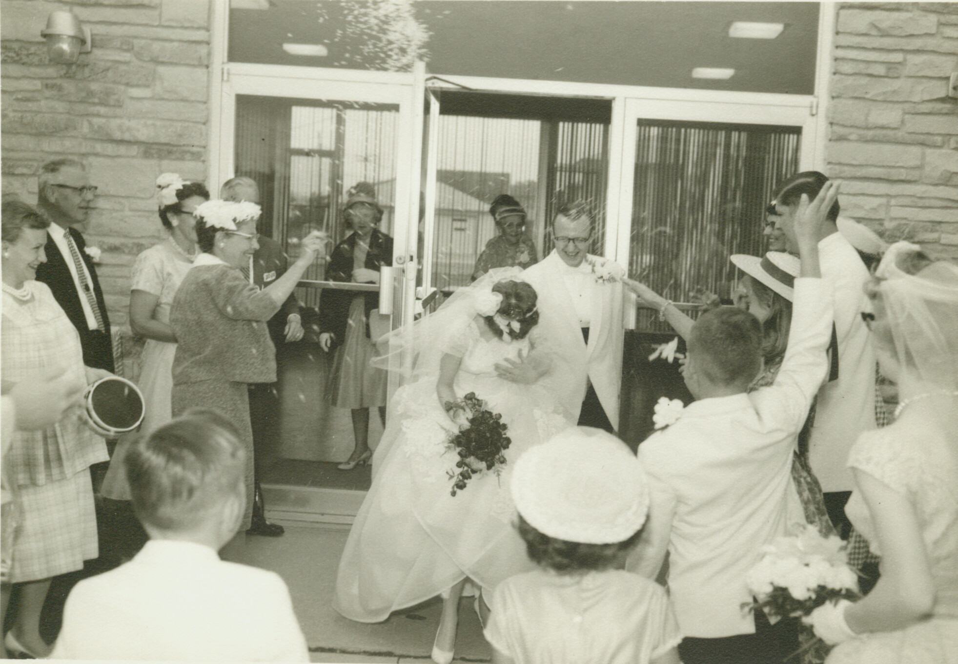 Bride in a wedding gown exits a building, surrounded by bridesmaids and guests throwing flowers.