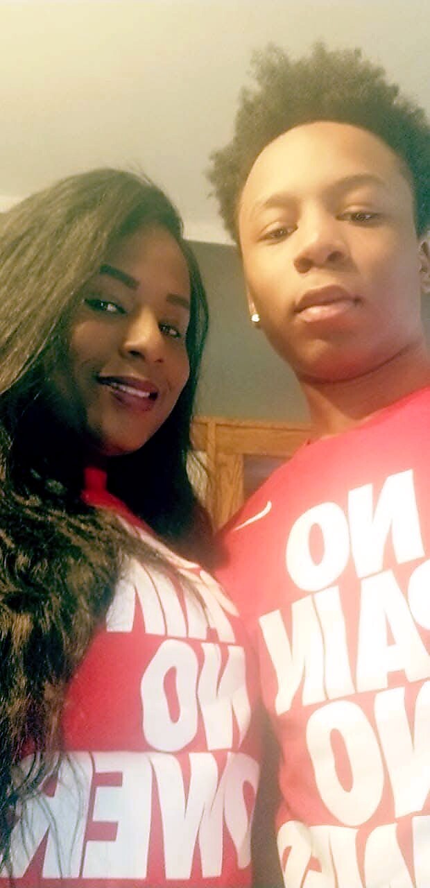 Two friends take a selfie indoors, wearing matching red shirts with large white text; the woman smiles at the camera while the man looks at the lens behind her.
