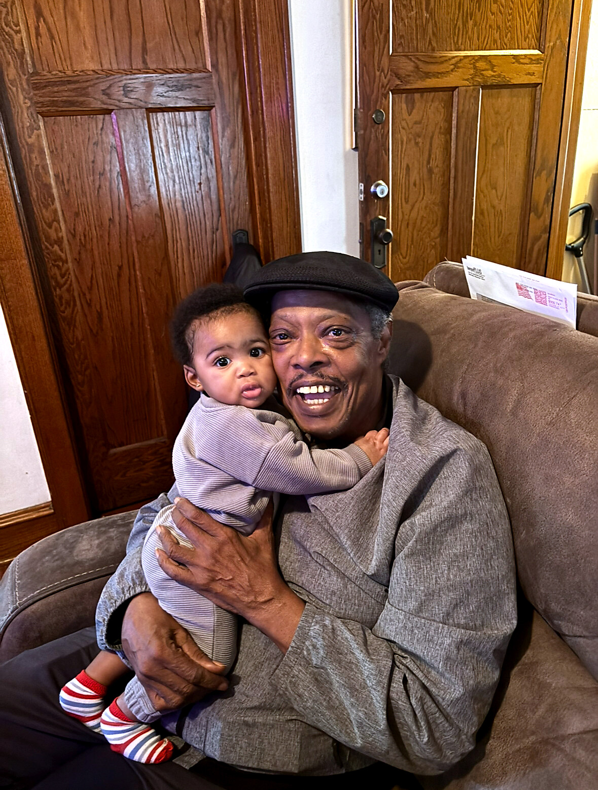 Older woman wearing a black hat smiles while holding a baby on her lap on a brown couch indoors, wooden doors in the background.