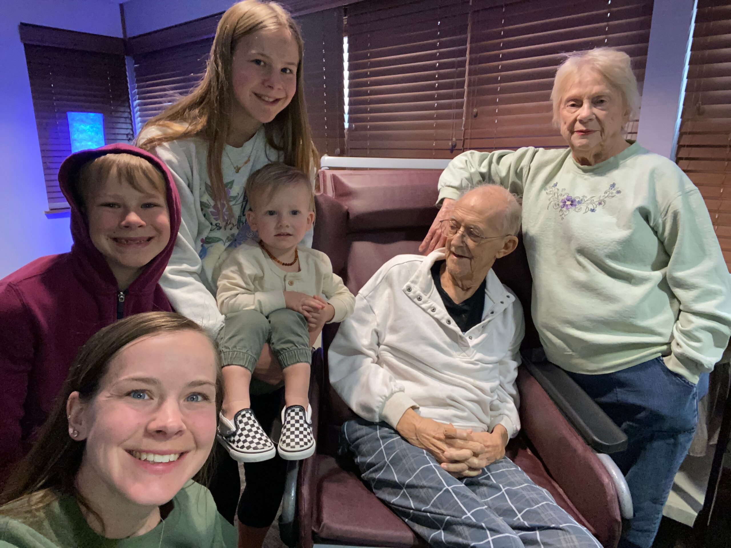Front-left woman takes a group selfie with two kids and two seniors posing on a maroon couch indoors among wooden blinds.