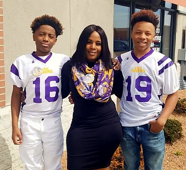 Three people pose outdoors: two LSU football players in white/purple jerseys (numbers 16 and 19) flank a woman in a black dress with an LSU scarf at the center.