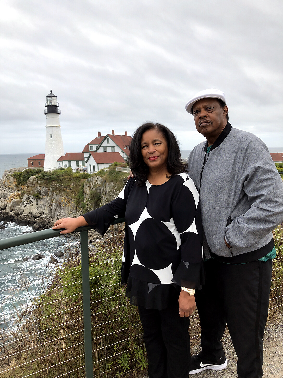 Couple standing at a coastal overlook with a white lighthouse and red-roofed houses behind them.