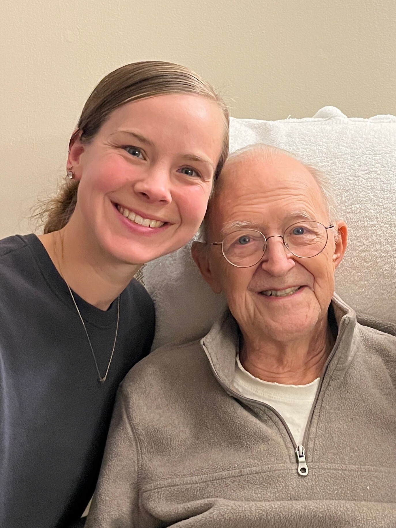 Young woman and elderly man smiling for a selfie indoors, seated on a beige couch.