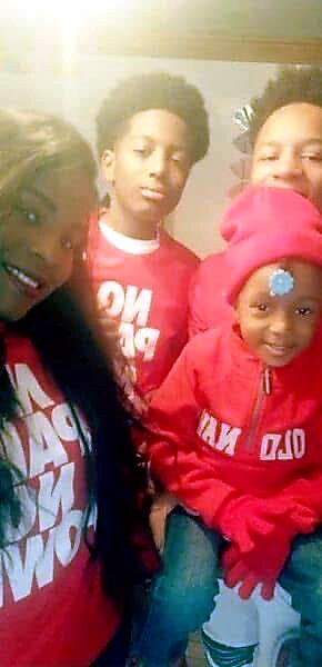 Four children pose for a photo indoors, wearing matching red hoodies and smiling at the camera, with sunlight in the background.